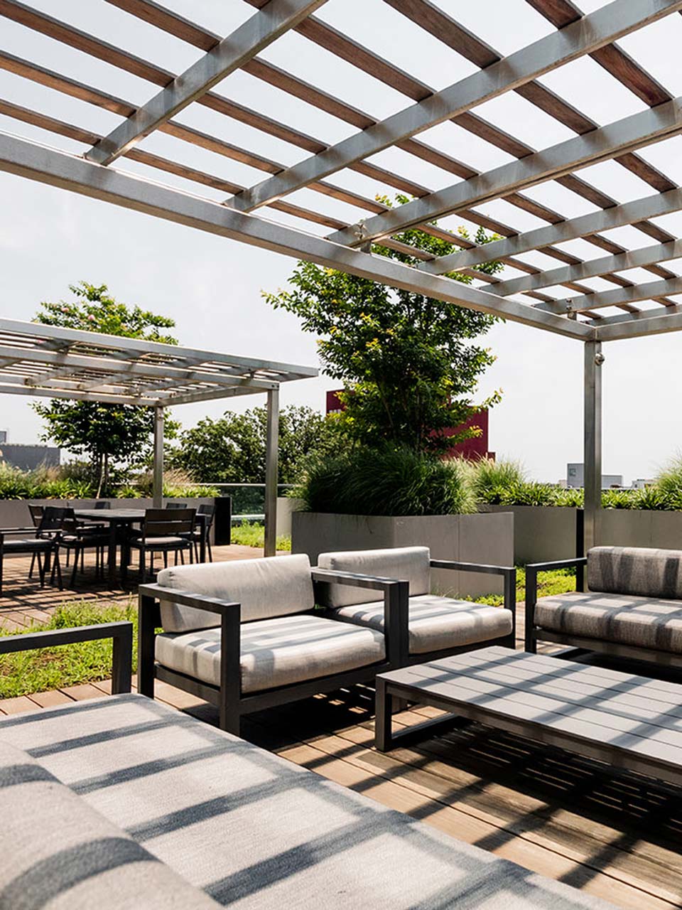 Modern rooftop terrace with black metal pergolas, gray upholstered lounge chairs, dining area in background, potted plants and ornamental grasses, geometric patterned pavers, and countryside views.
