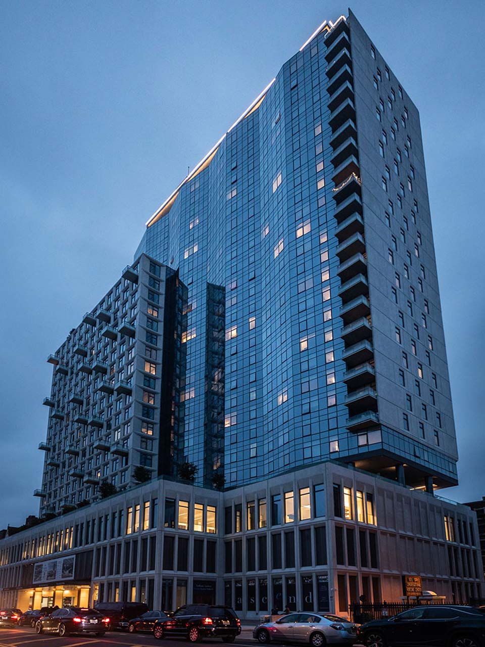 A modern mixed-use high-rise building photographed at dusk, featuring a glass and steel tower with illuminated windows rising above a contemporary ground-level retail base with large windows, with parked cars visible in the foreground against a twilight blue sky.