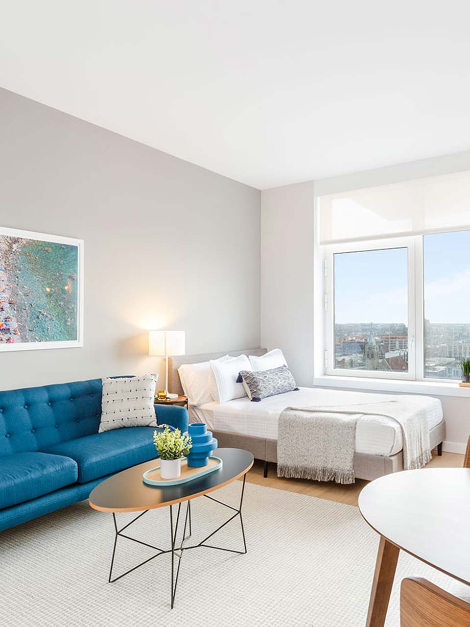 A modern studio apartment with a blue tufted sofa, mid-century coffee table, queen bed with grey headboard, floor-to-ceiling windows overlooking a city skyline, white walls, and minimalist decor accents.