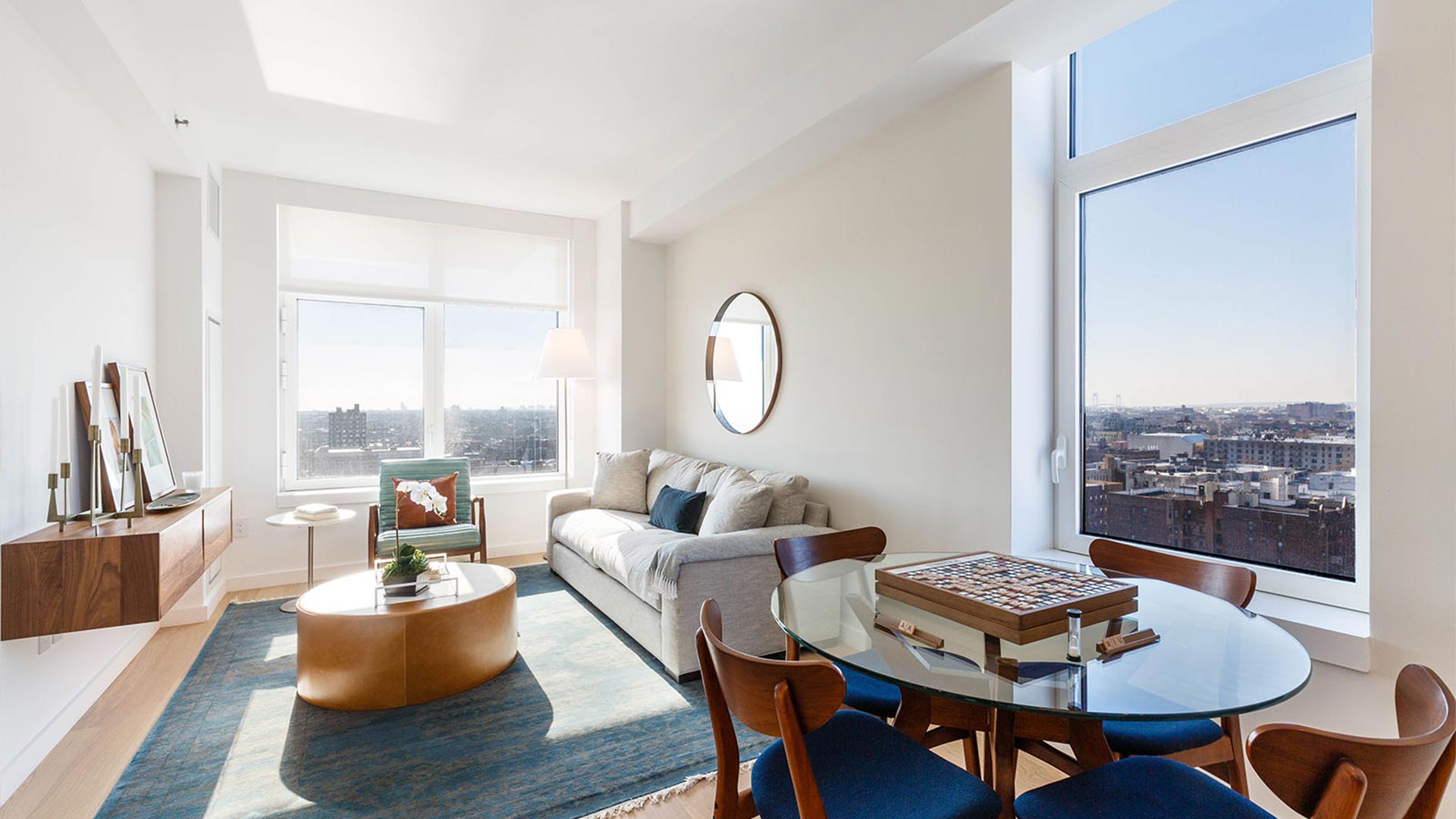 A modern high-rise apartment living room with floor-to-ceiling windows overlooking a city skyline, featuring mid-century furniture including a beige sofa, wooden dining table with blue chairs, round gold coffee table, and light wood cabinetry.