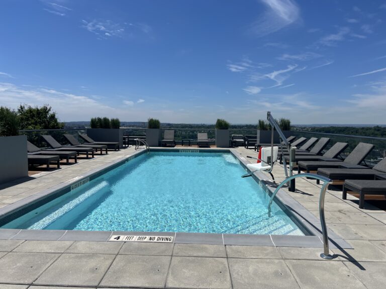 A rooftop swimming pool with crystal-clear turquoise water, surrounded by gray loungers and planters, with a metal diving board on the right side. The pool deck features depth markers and 